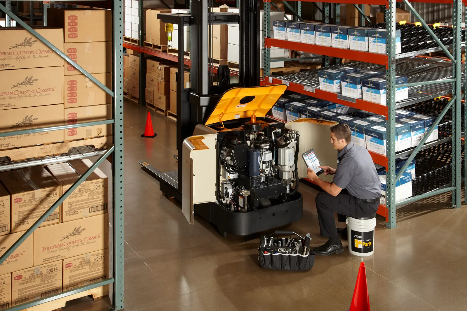 Technician recording a scheduled inspection beside an open Crown truck in a warehouse aisle